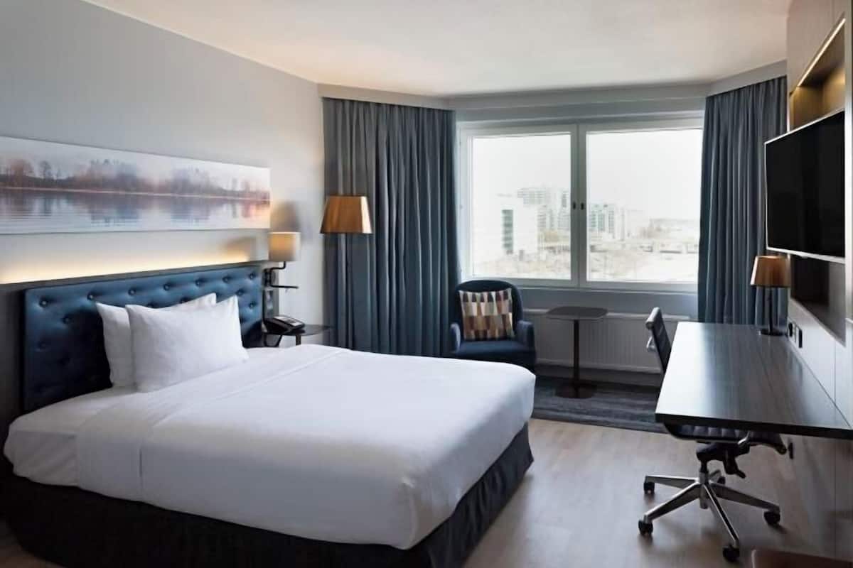A hotel room with a large bed, a desk, a chair, a TV, and a window with a city view.