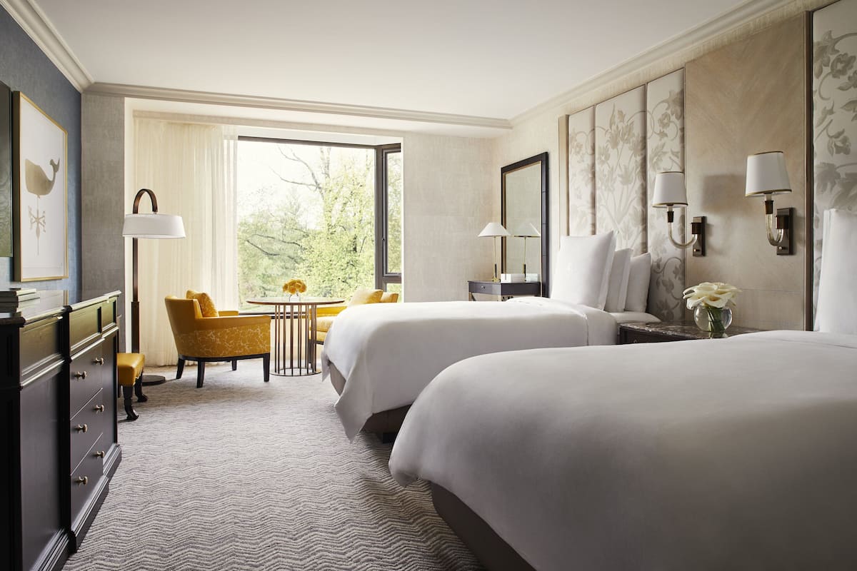 Ein Hotelzimmer mit zwei Betten, einem Schreibtisch, einem Stuhl, einem Schminktisch und einem großen Fenster mit Blick auf Bäume.