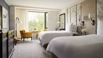 A hotel room with two beds, a desk, a chair, a dresser, and a large window with a view of trees.