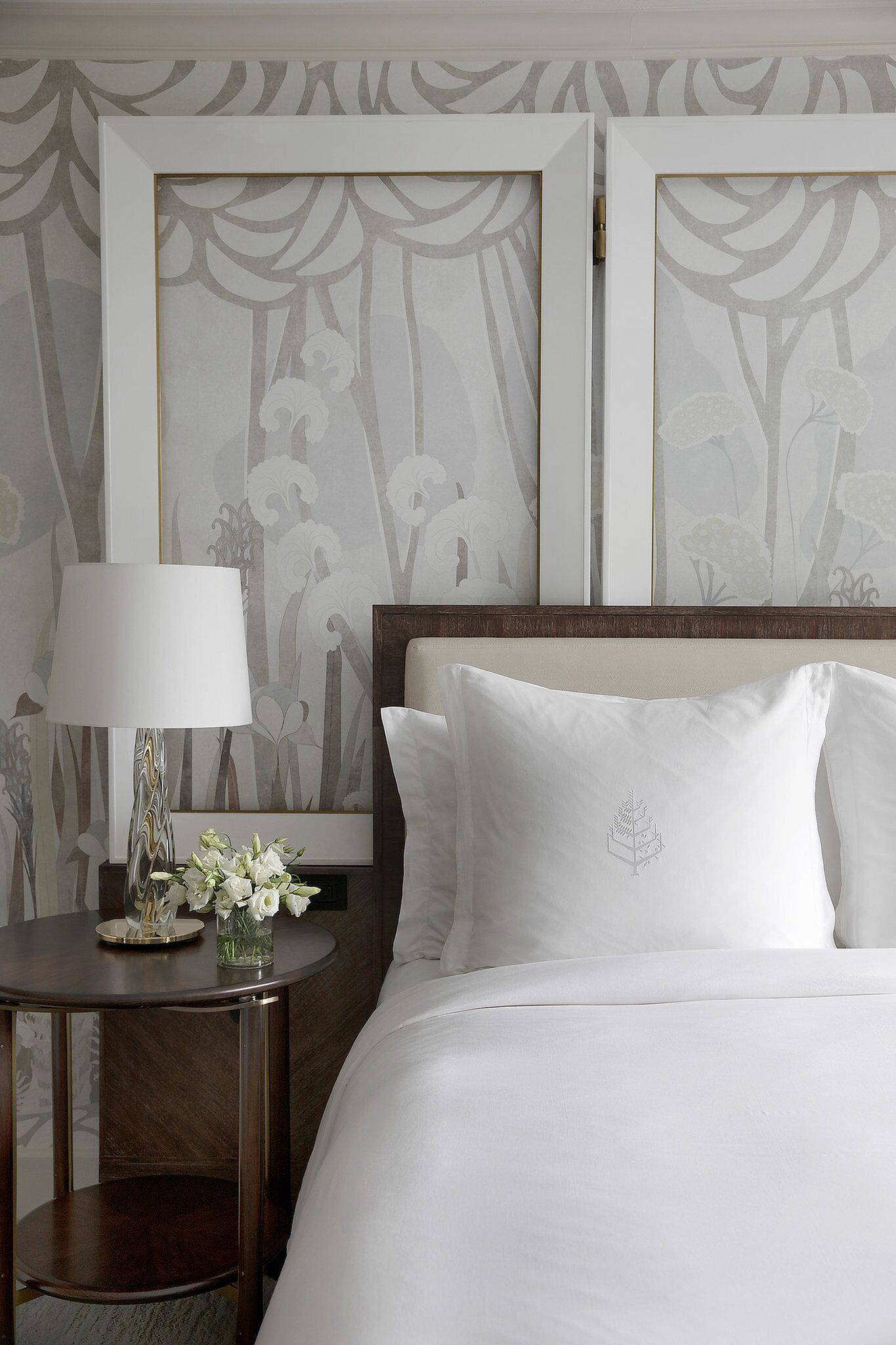 Een net opgemaakt bed met witte lakens, een nachtkastje met een lamp en een bloemenvaas, en een wand met ingelijste kunstwerken.