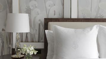 Een net opgemaakt bed met witte lakens, een nachtkastje met een lamp en een bloemenvaas, en een wand met ingelijste kunstwerken.