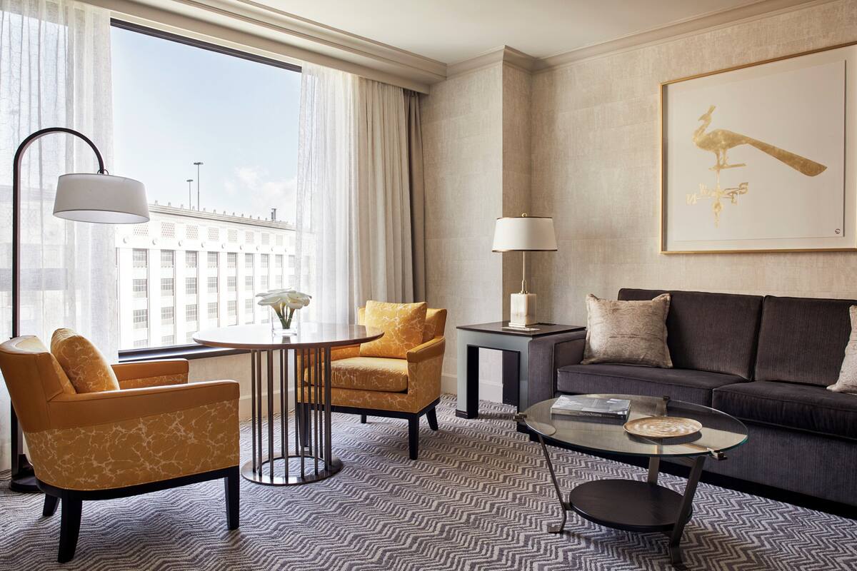 Ein modernes Hotelzimmer mit einer Couch, Sesseln, einem Couchtisch und einem großen Fenster mit Blick auf die Stadt.