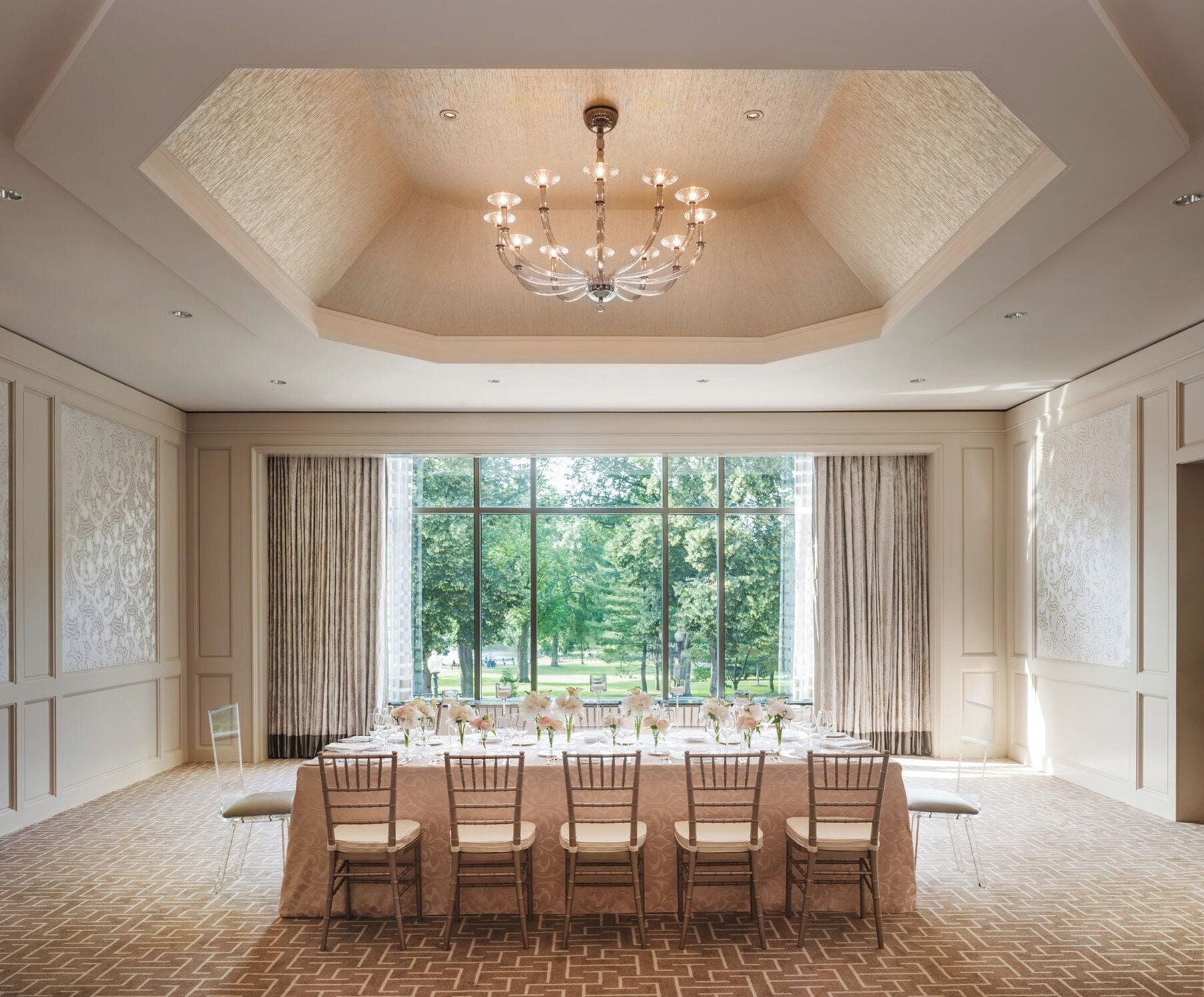 A formal dining room with a large table set for six, a chandelier, and a view of a garden through large windows.