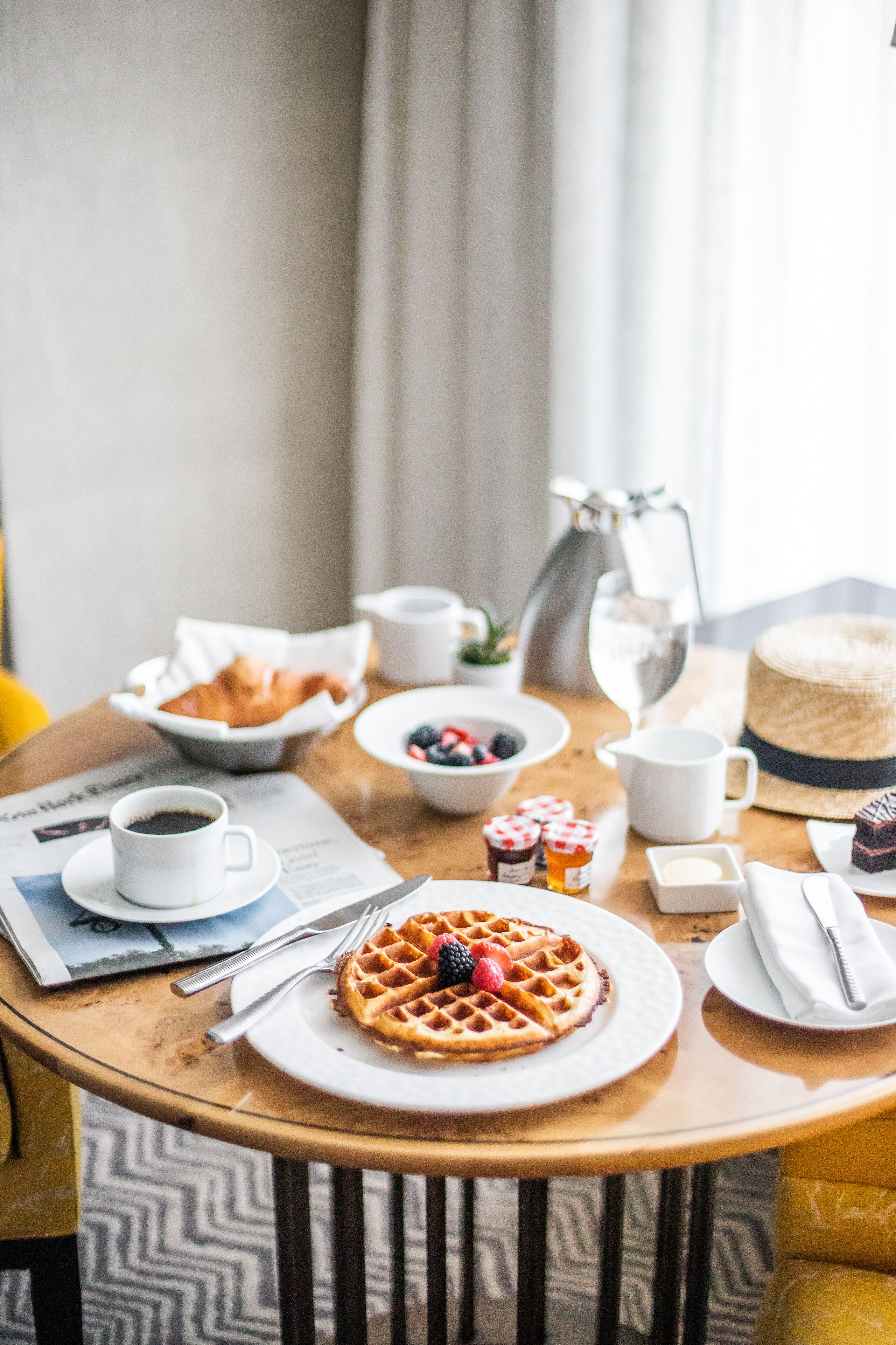 A breakfast table with waffles, a cup of coffee, a bowl of fruit, and a cake.