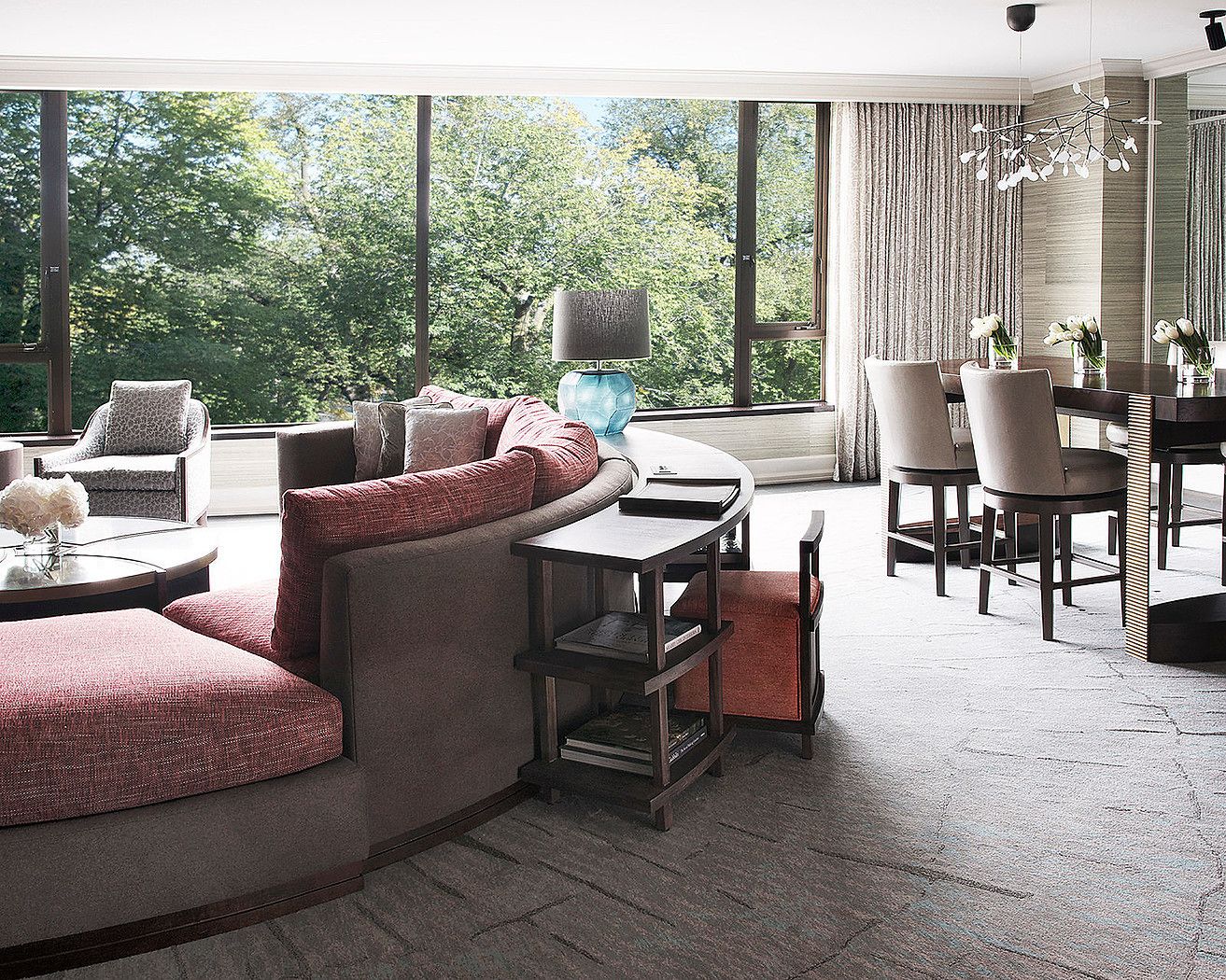 A modern living room with a curved sofa, a dining table with chairs, and a large window offering a view of trees.