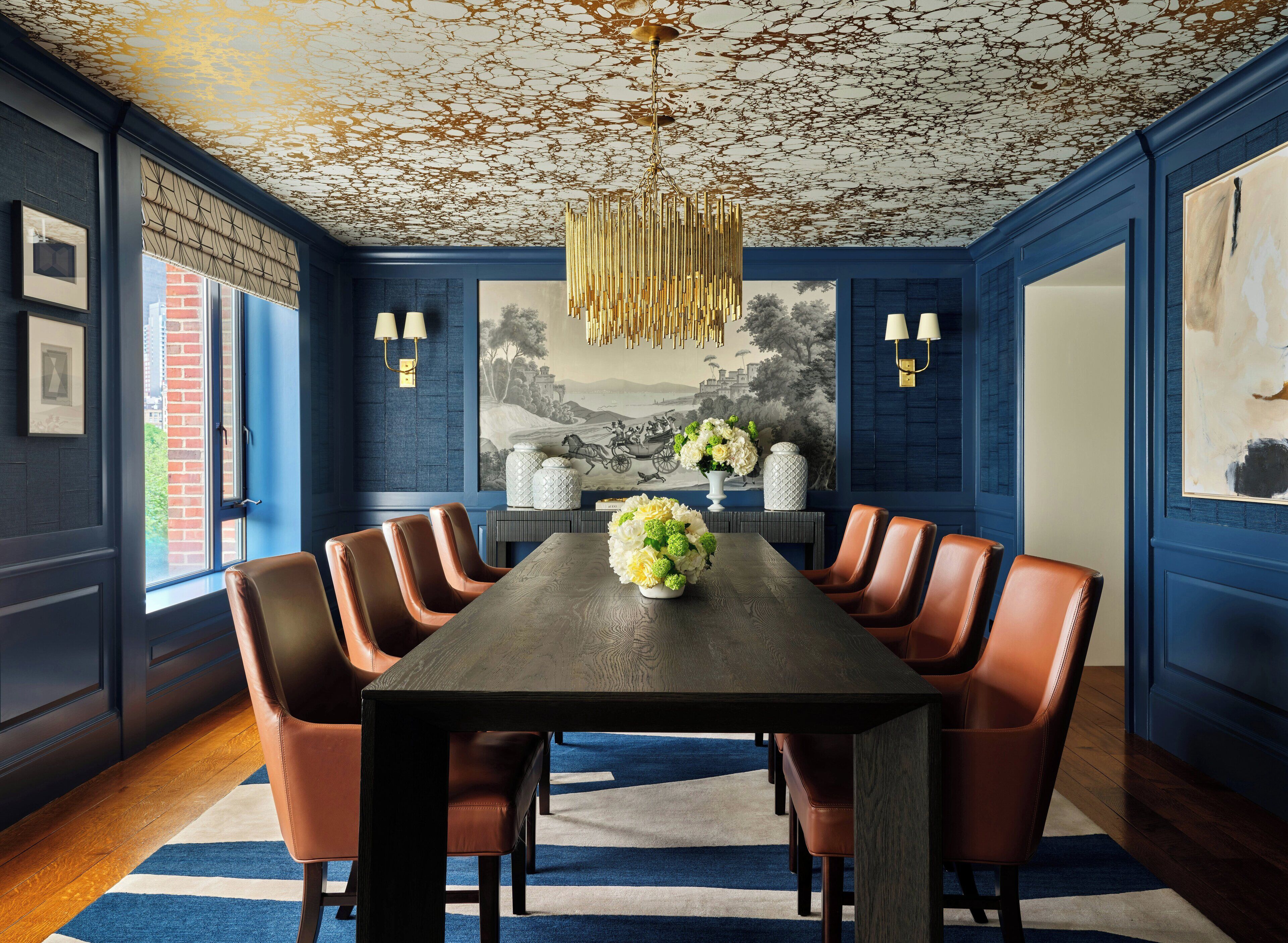 Comedor con una mesa larga, seis sillas de cuero marrón, una araña y una alfombra con estampado azul y blanco.