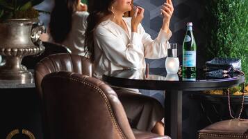 Een vrouw zittend aan een tafel met een drankje, omringd door stoelen en een potplant.