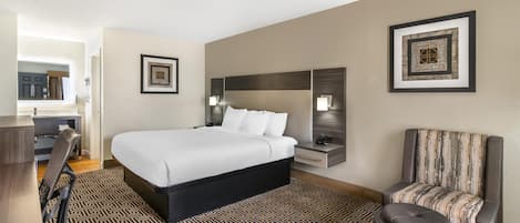A hotel room with a large bed, a desk, a chair, and a small stool. There are framed pictures on the walls and a lamp on the desk.