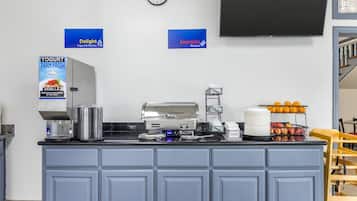 A kitchen counter with a yogurt dispenser, coffee maker, and fruit. There are signs for 'Delight Yogurt & Pastries' and 'Energize Proteins' on the wall.