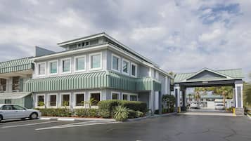 A two-story hotel with a green roof and white exterior. There is a covered entrance with a sign, and a parking lot with cars.