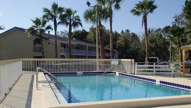 A swimming pool with a white deck, surrounded by palm trees and a building with balconies. - Rodeway Inn Leesburg Chain of Lakes (Leesburg)