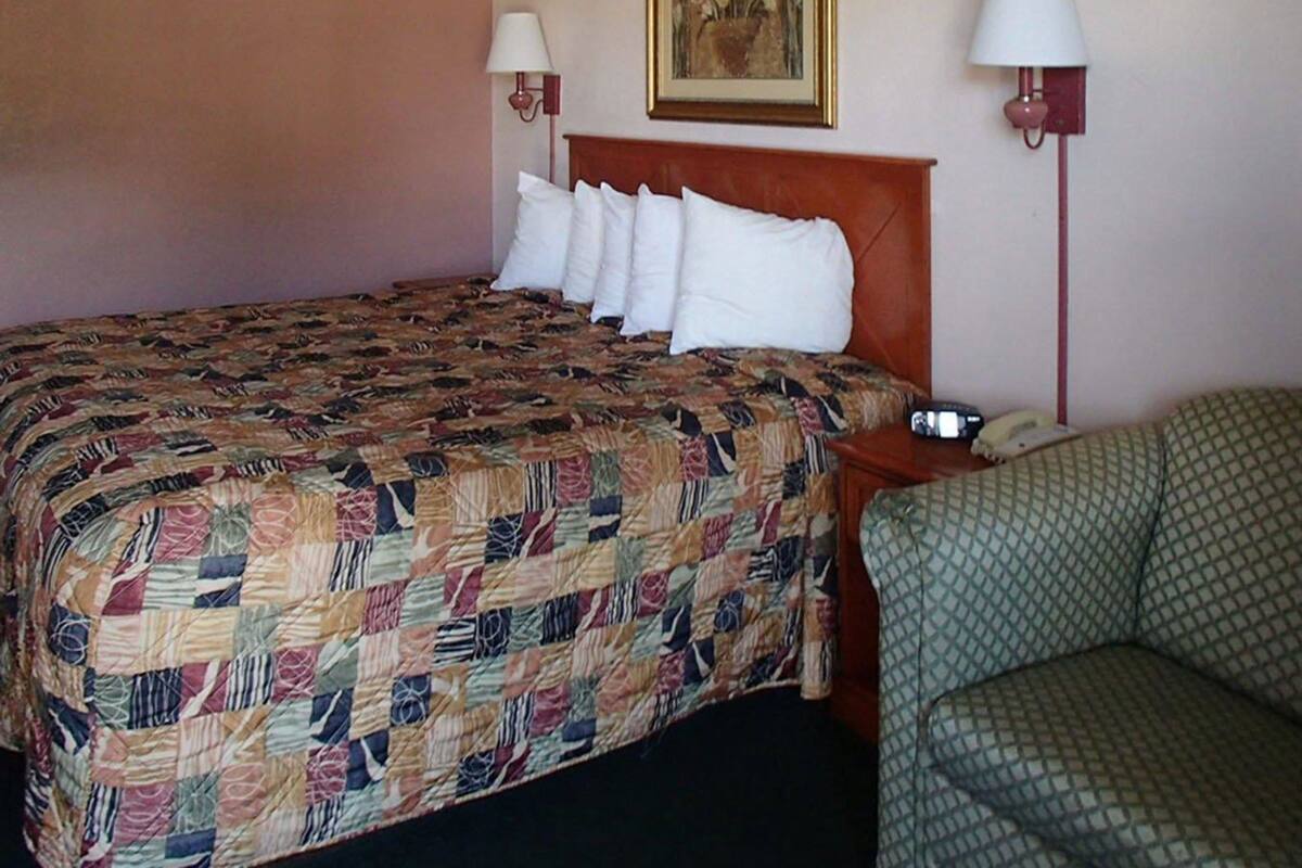 Une chambre d’hôtel avec un lit, une chaise, deux lampes de chevet, un tableau au mur et un téléphone sur la table de chevet.