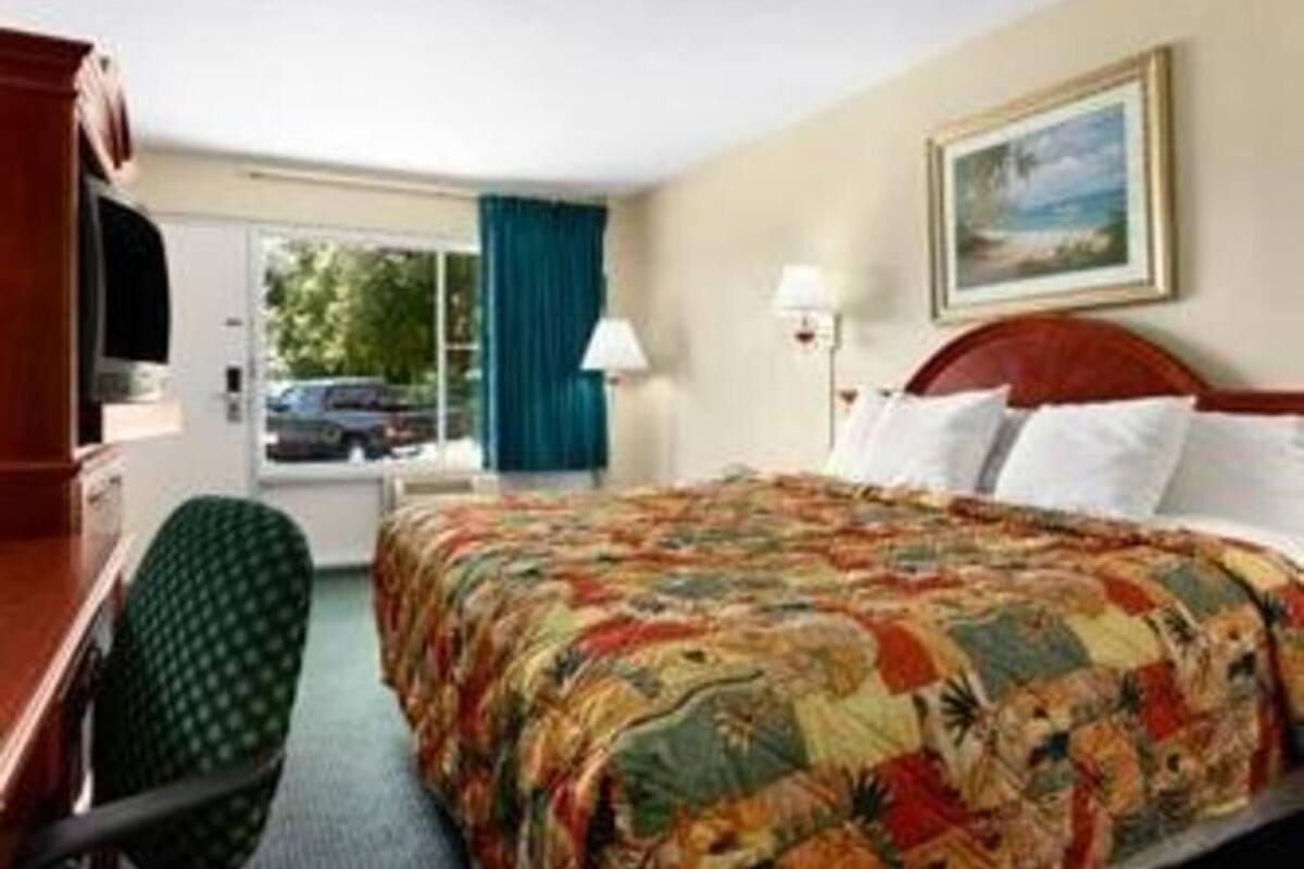 Une chambre d’hôtel avec un lit, un bureau, une chaise, une fenêtre avec des rideaux, un tableau au mur et une lampe.