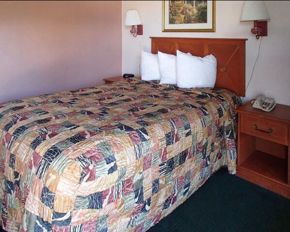 A neatly made bed with a patterned quilt, a wooden headboard, two bedside lamps, a bedside telephone, and a small wooden nightstand. - Rodeway Inn Leesburg Chain of Lakes (Leesburg)