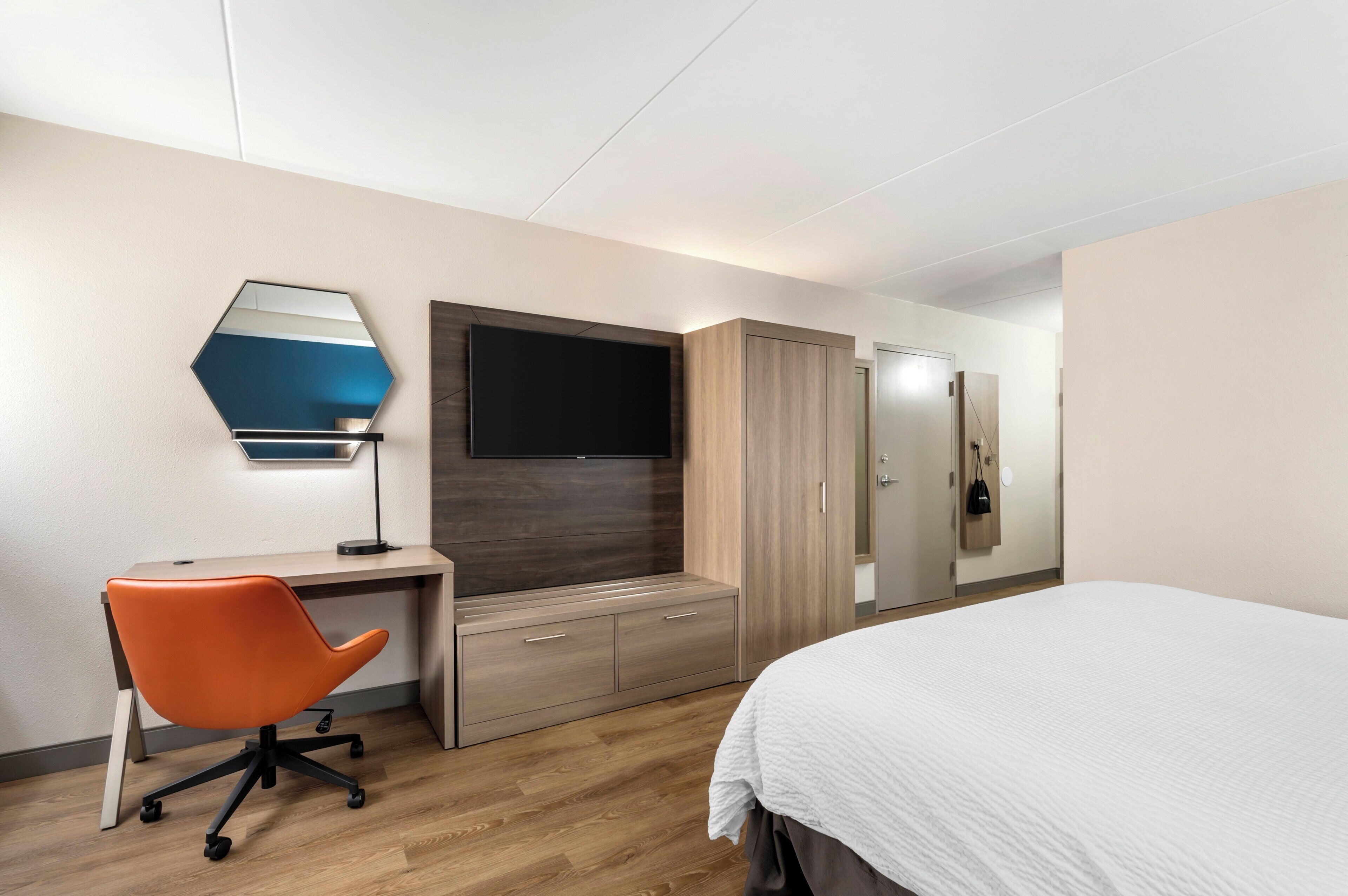 Una habitación de hotel con una cama, un escritorio con una silla naranja, un televisor, un armario y un espejo.