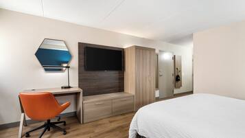 Una habitación de hotel con una cama, un escritorio con una silla naranja, un televisor, un armario y un espejo.