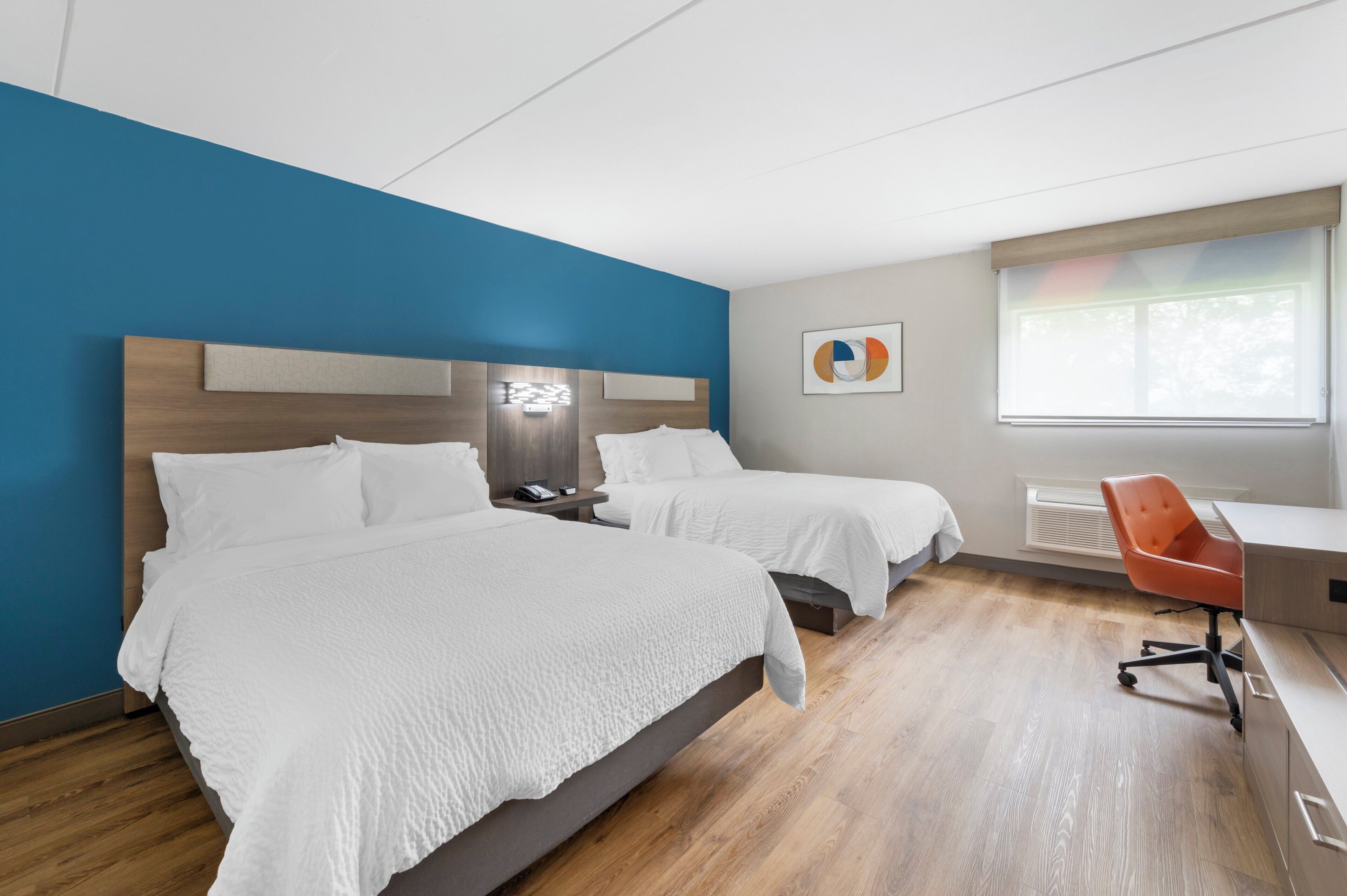 A hotel room with two beds, a desk, a chair, a window, and a wall art piece.