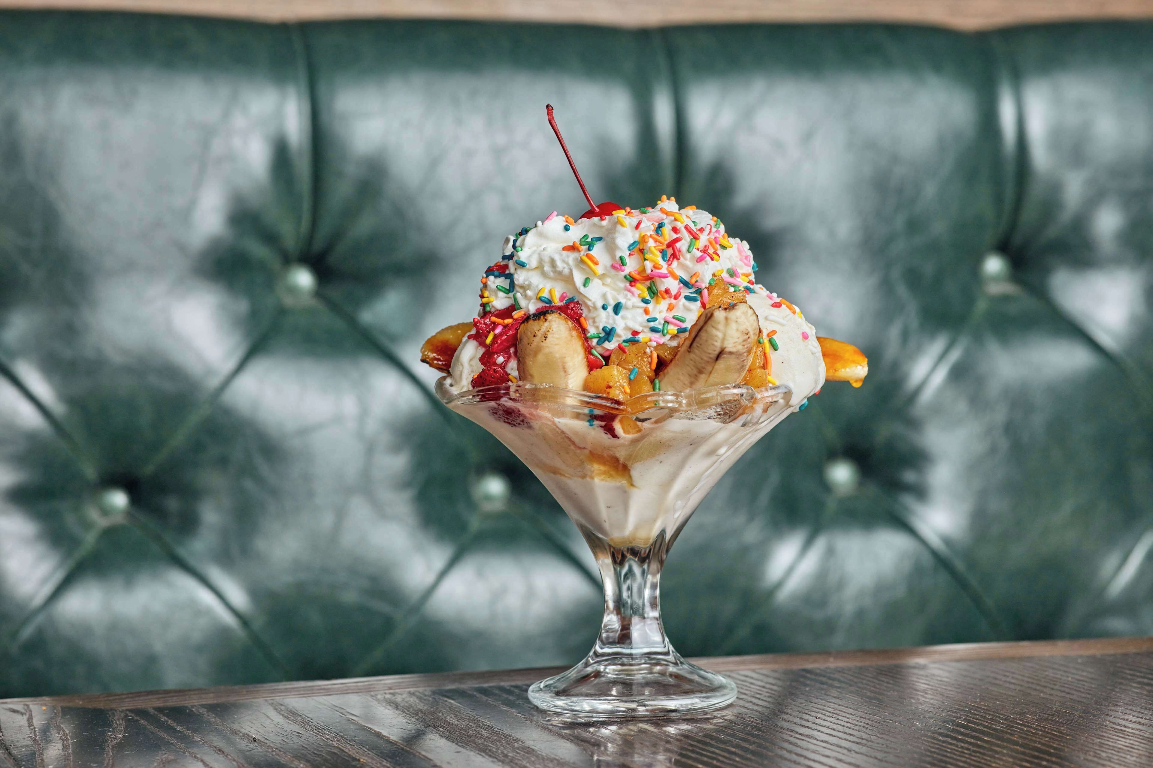 A dessert in a glass bowl on a wooden table with a green upholstered background.