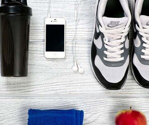 Une paire de baskets Nike, un téléphone intelligent, une bouteille d’eau, une paire d’écouteurs, une serviette bleue et une pomme, le tout posé sur une surface en bois.