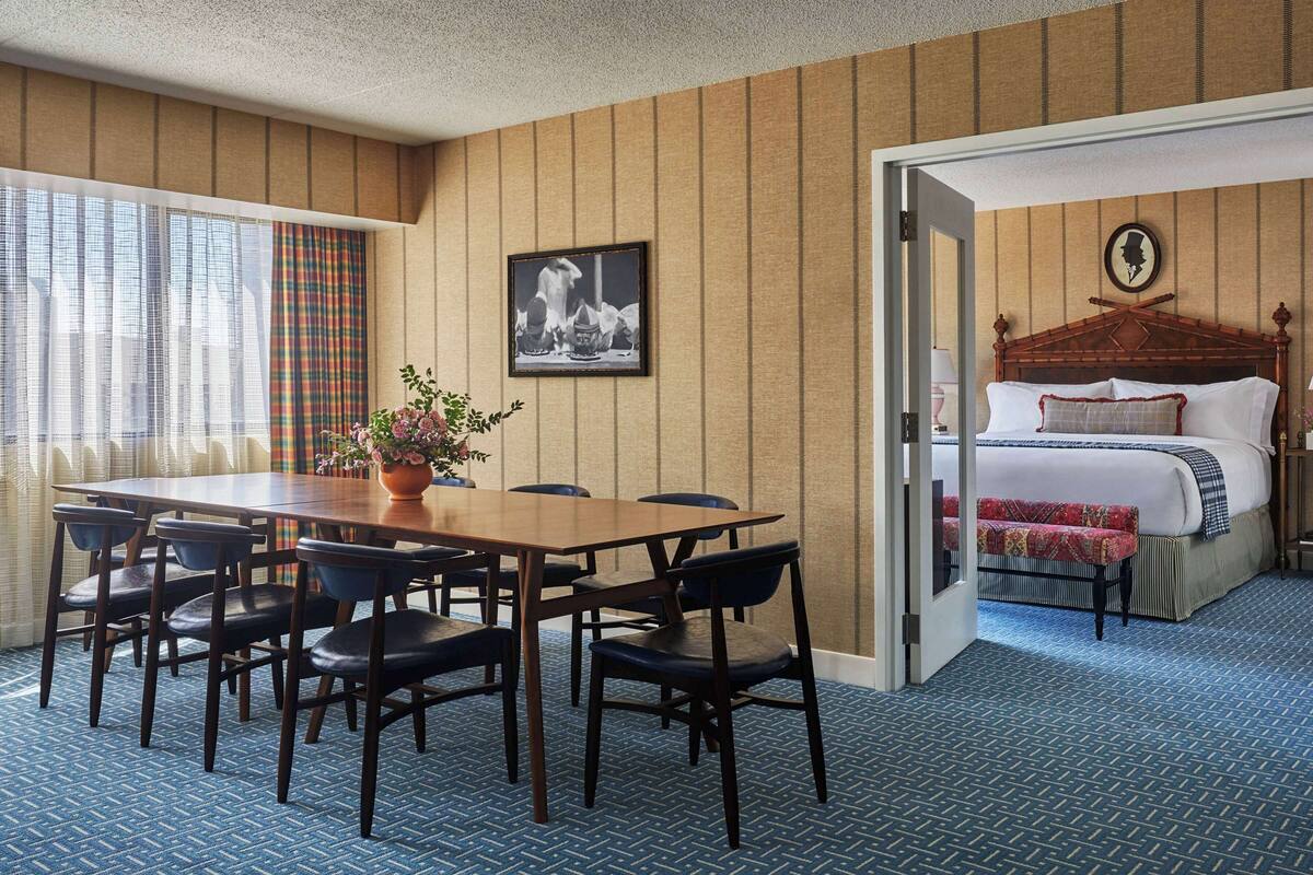 A hotel room with a wooden dining table, chairs, a potted plant, a bed with a headboard, and a nightstand with a lamp.