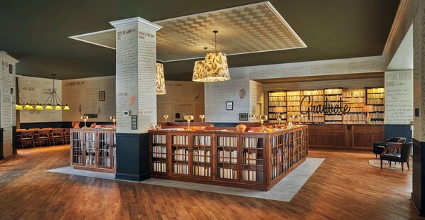 Une bibliothèque avec des étagères en bois, un espace de lecture central doté de fauteuils et un plafond orné d’un motif géométrique.