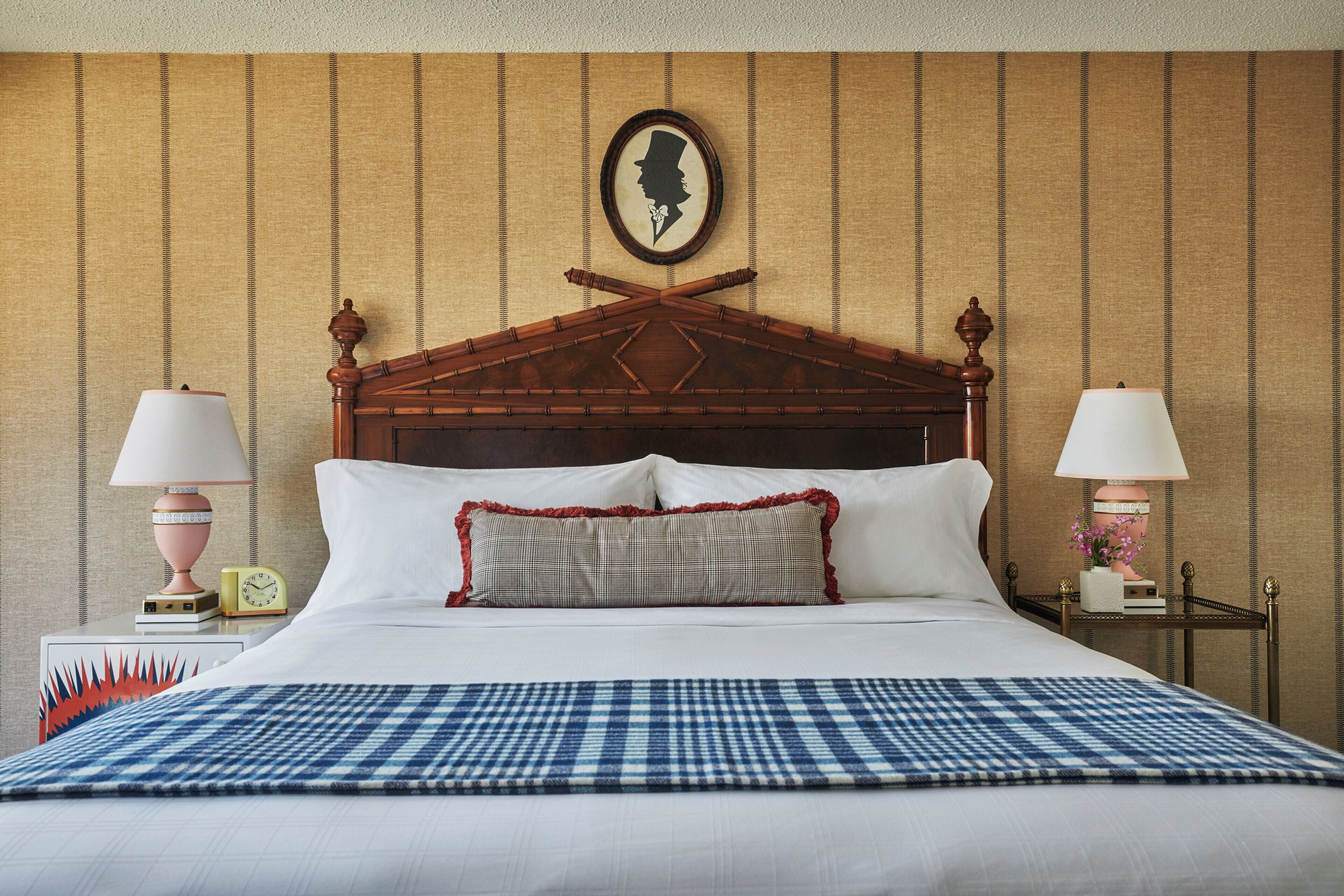 A neatly made bed with a checkered bedspread, two bedside tables with lamps, a wall clock, and a framed silhouette on the wall.