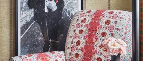 A room with a floral-patterned armchair, a framed artwork on the wall, and a table with a vase of flowers.