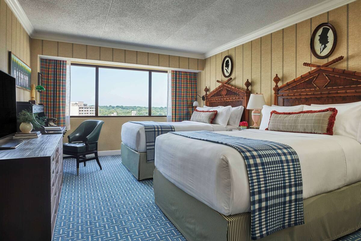 A hotel room with two beds, a desk with a chair, a television, and a large window offering a city view.