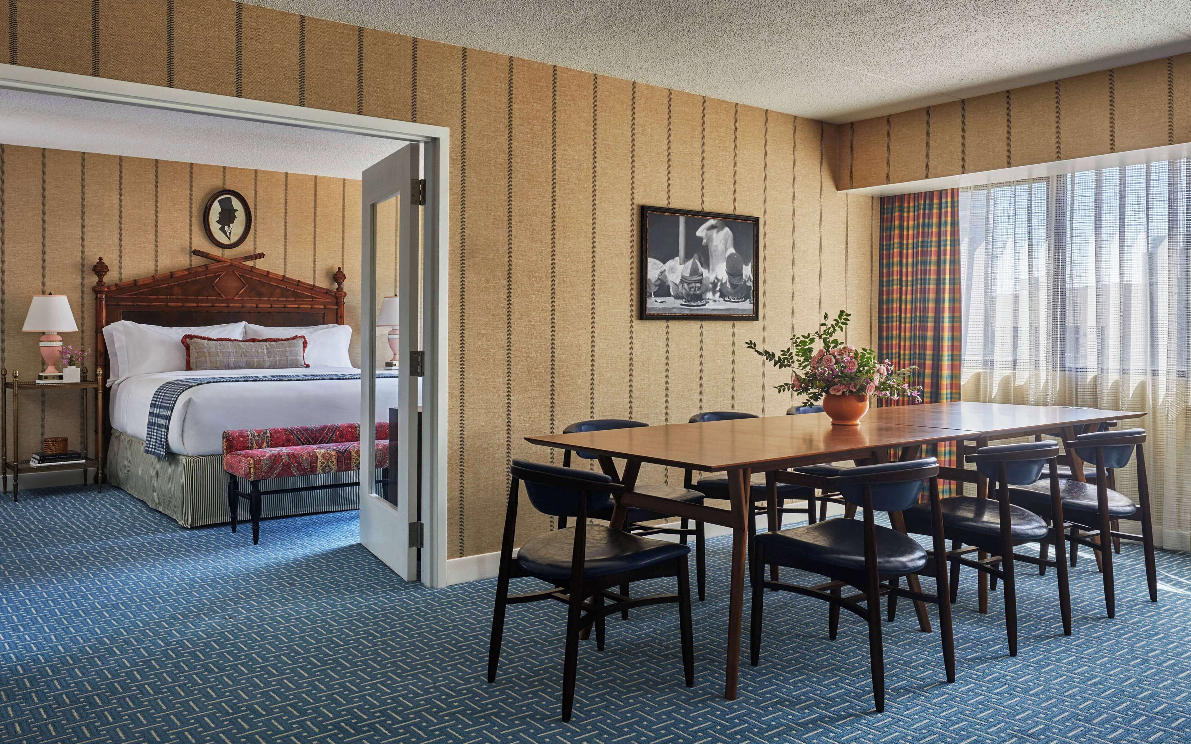 A hotel room with a bed, a dining table with chairs, a potted plant, and a framed picture on the wall.