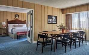 A hotel room with a bed, a dining table with chairs, a potted plant, and a framed picture on the wall.