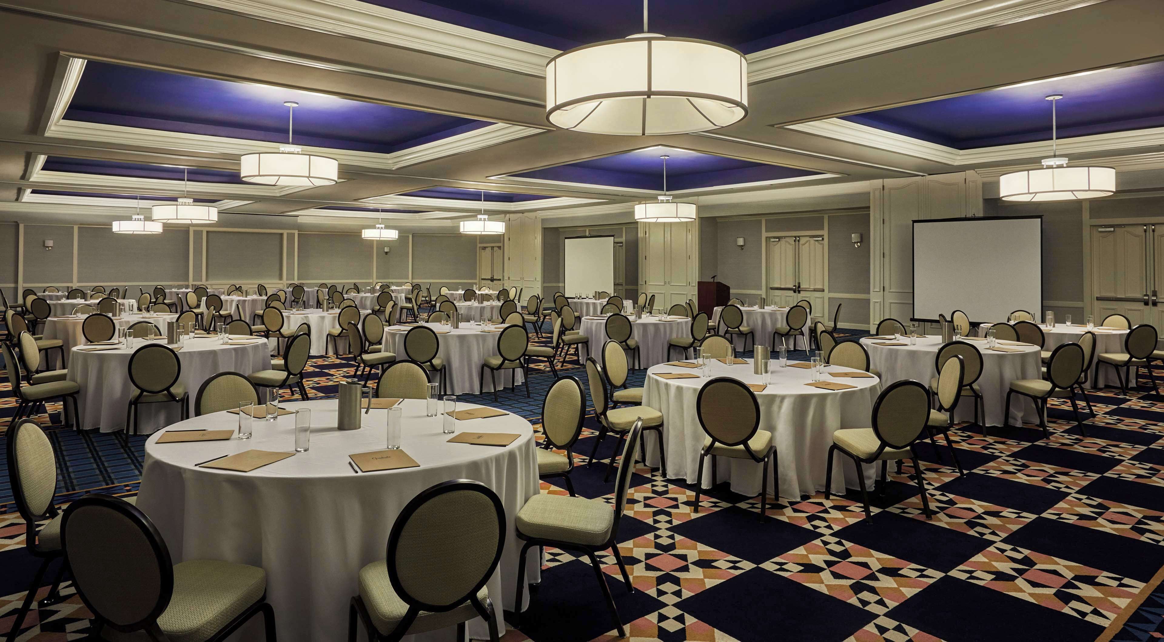A large conference room with round tables and chairs, a patterned carpet, and ceiling chandeliers.