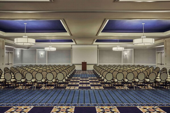 Un salón de conferencias con hileras de sillas, alfombra estampada e iluminación colgante.