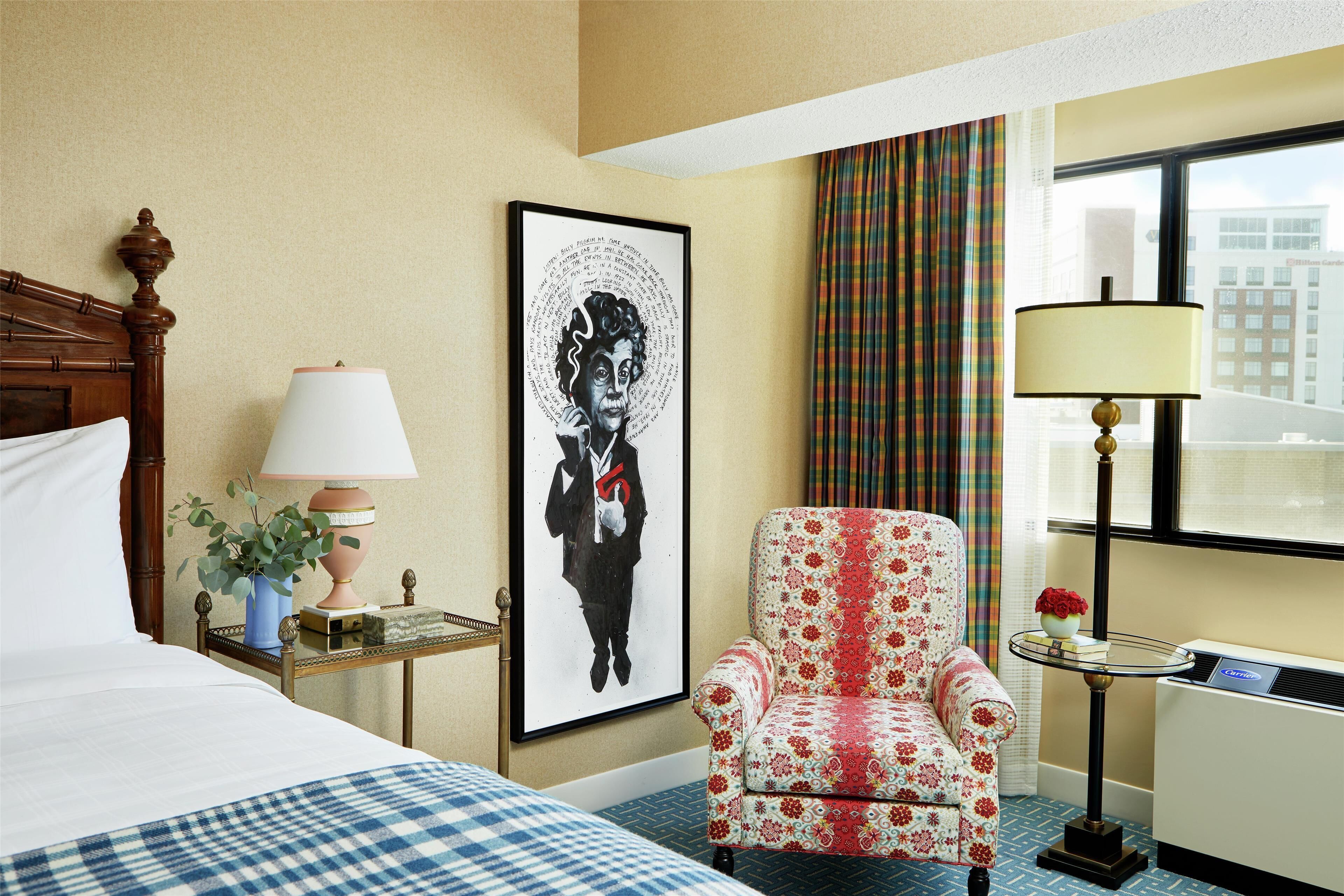 A hotel room with a bed, a chair, a table with a lamp, a framed picture on the wall, and a window with curtains.