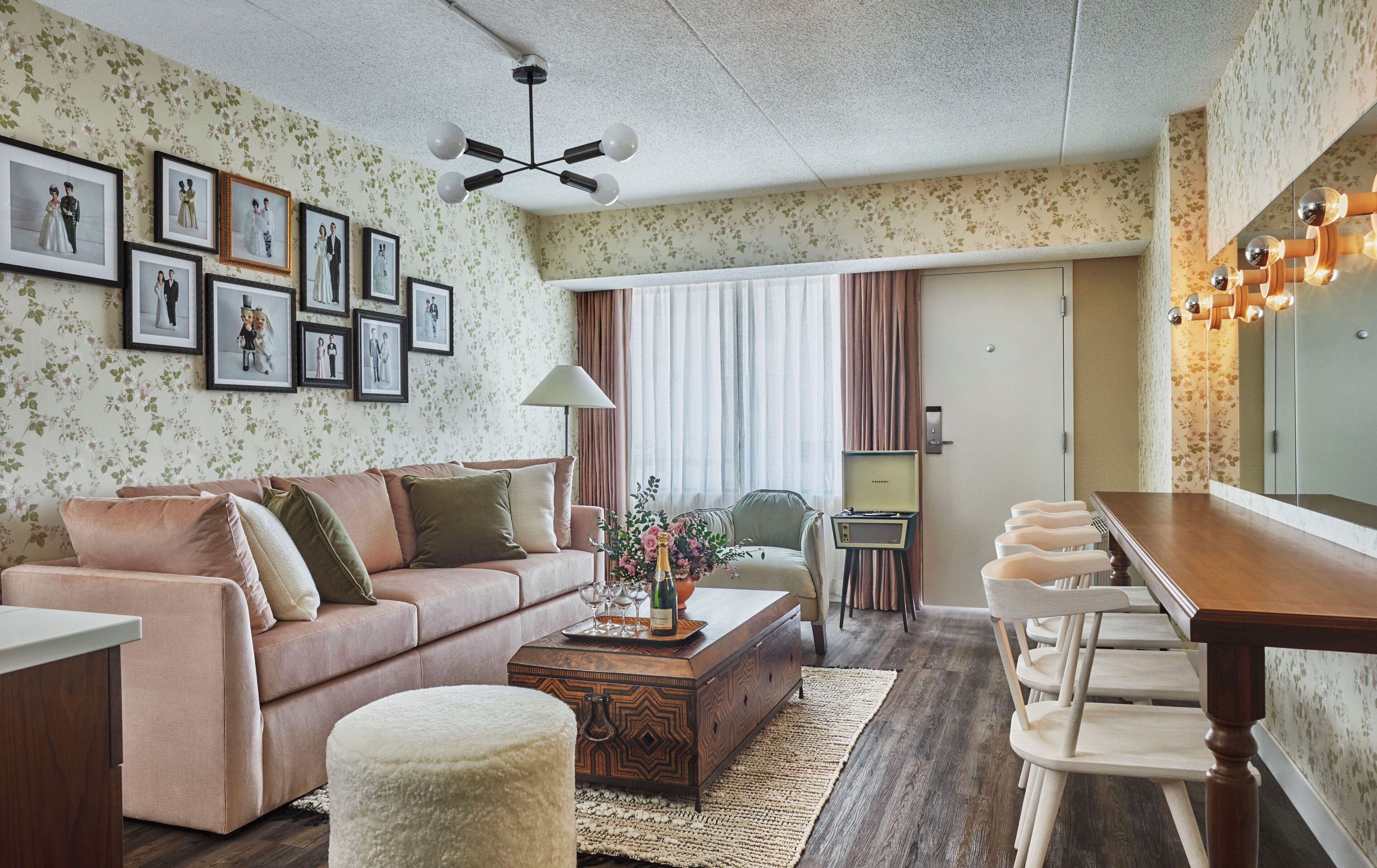 Een woonkamer met een bank, poef, salontafel, en een eethoek met een tafel en stoelen. Er hangen ingelijste foto’s aan de muur en er is een kroonluchter.