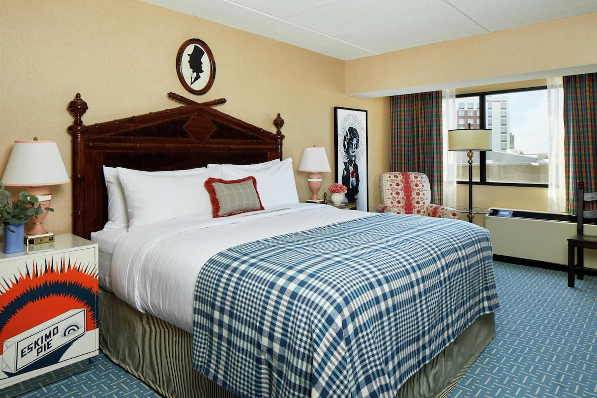 A hotel room with a large bed, two bedside tables with lamps, a chair, a framed picture on the wall, and a window with curtains.