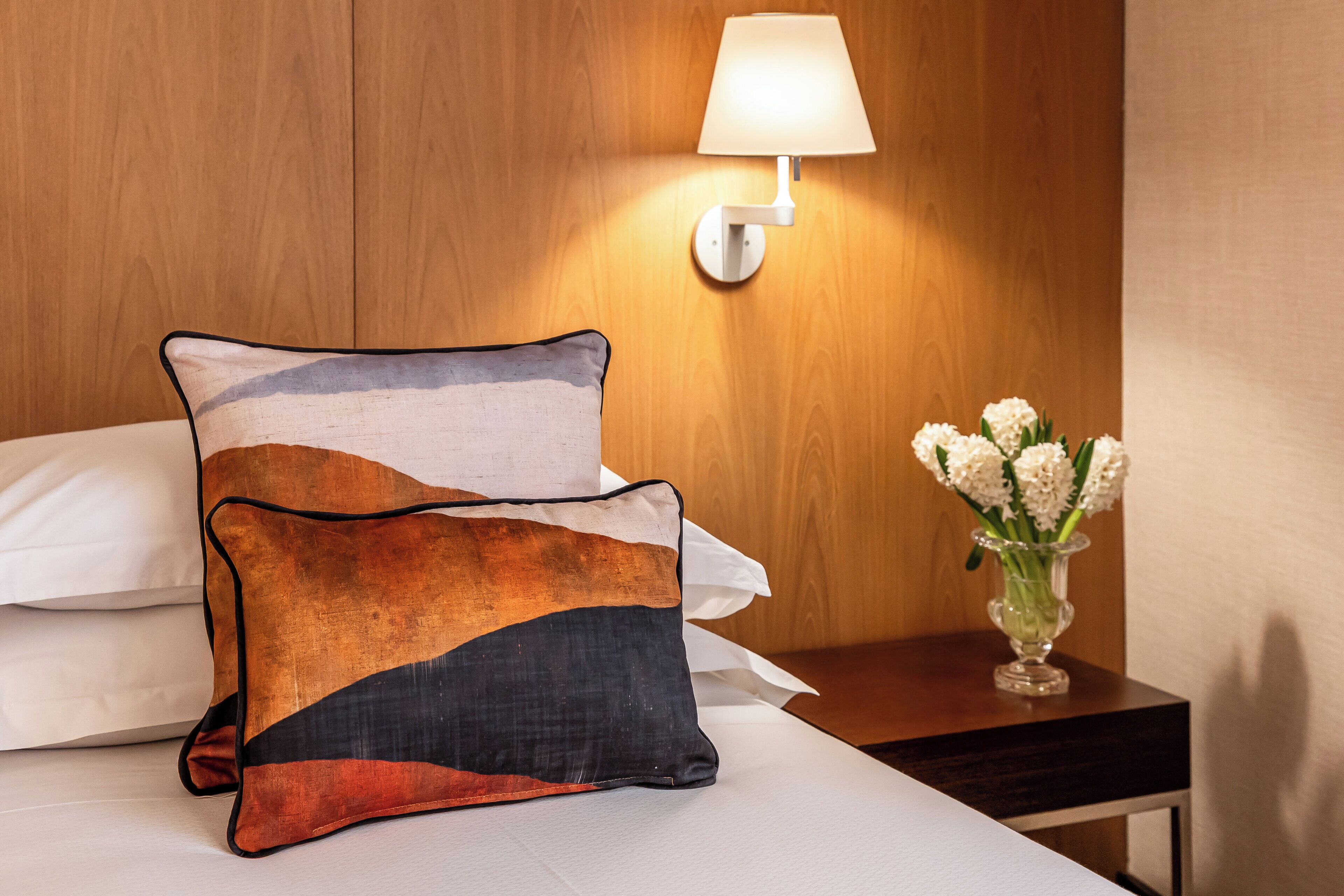 A bed with two pillows, a nightstand with a vase of flowers, and a wall-mounted lamp.