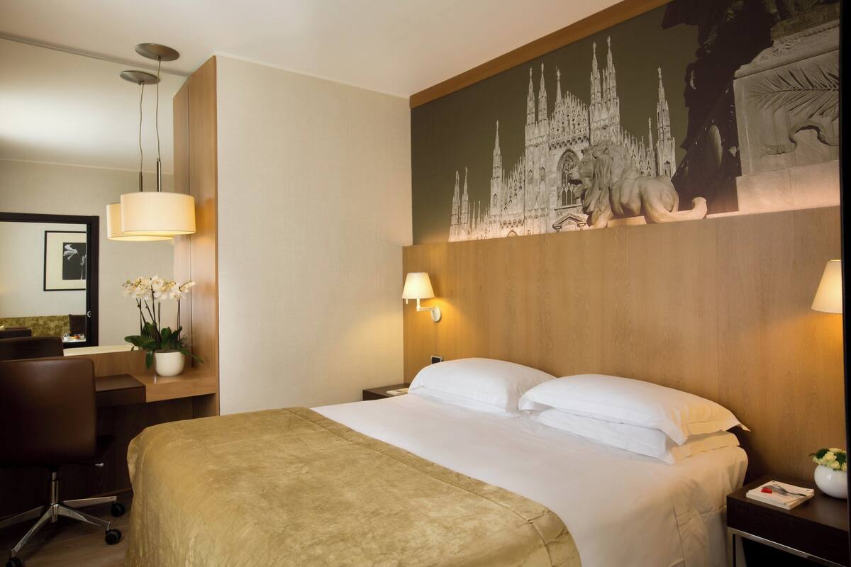 A hotel room with a large bed, a desk with a chair, a lamp, and a wall-mounted artwork of a castle.