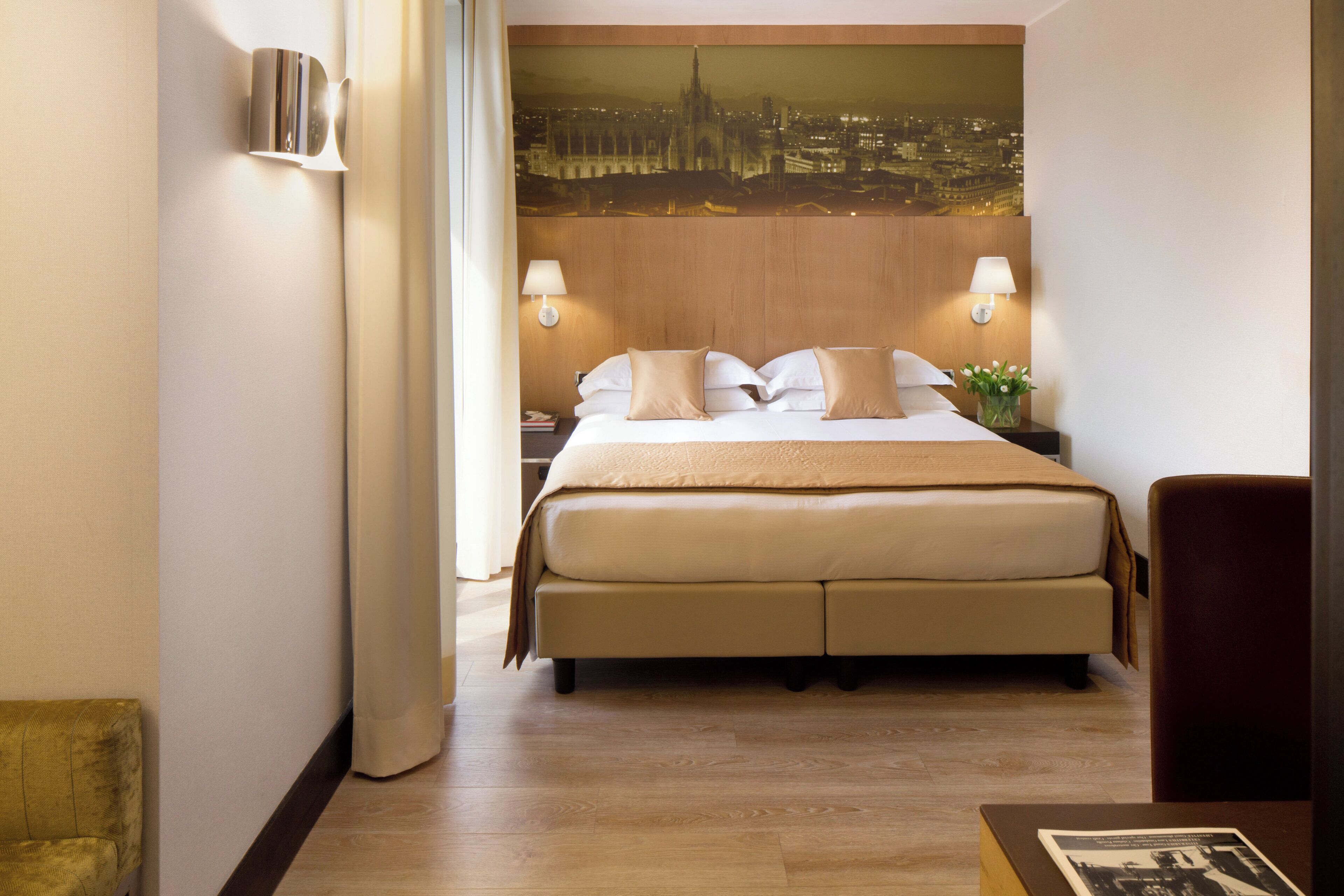 A hotel room with a large bed, two bedside lamps, a nightstand, a wooden headboard, and a cityscape painting on the wall.