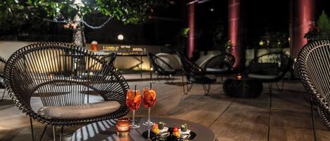 A well-lit outdoor seating area with wicker chairs and a round table, featuring a drink and snacks.