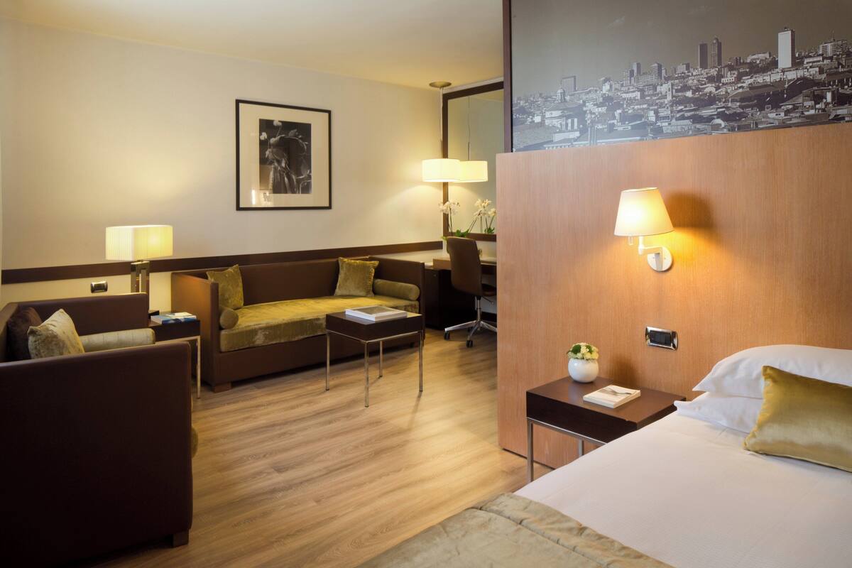 A hotel room with a bed, a sofa, a desk, a chair, and a cityscape view.