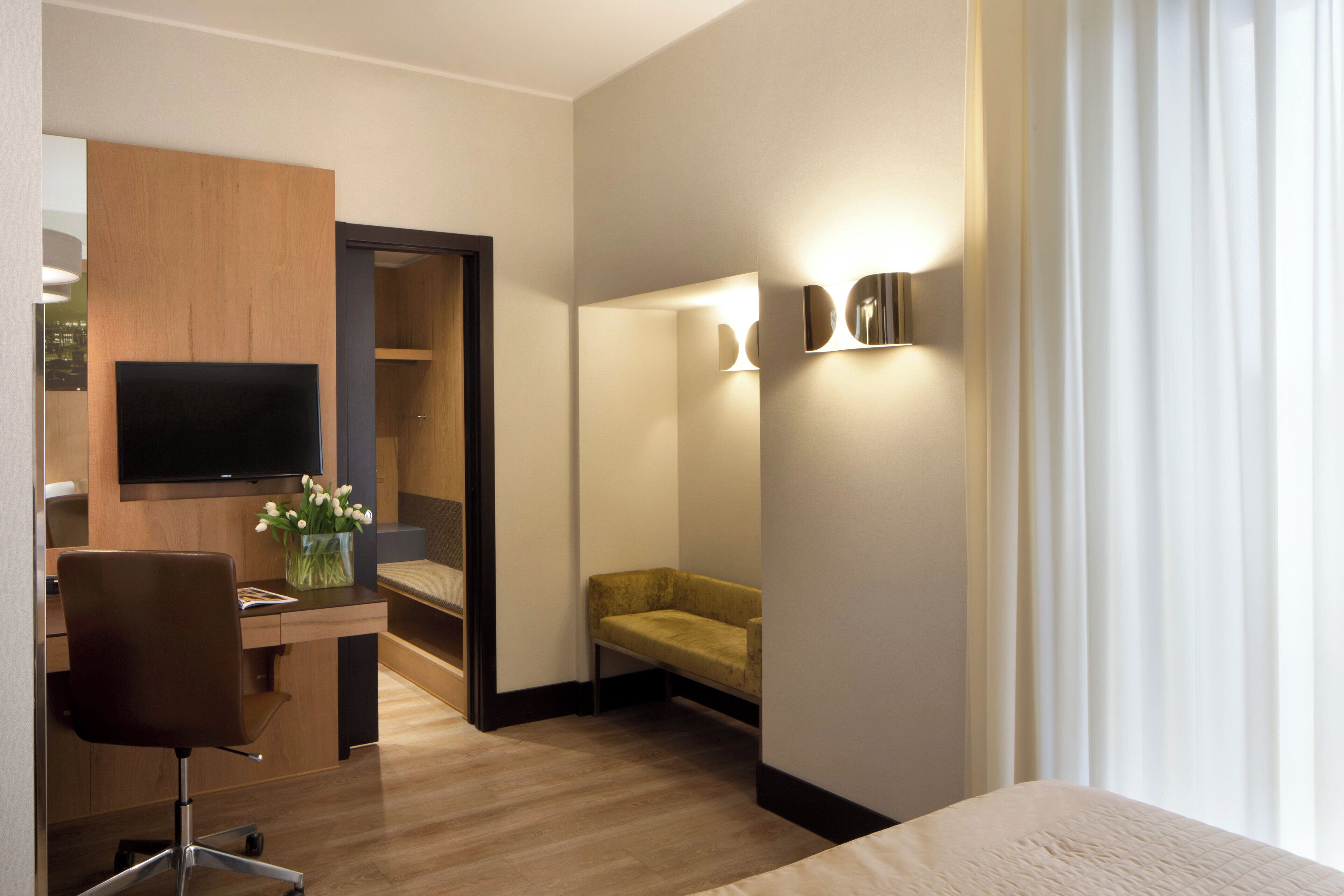 Una moderna camera d'hotel con una scrivania in legno, una TV a schermo piatto, un divano confortevole e un'ampia finestra con tende.