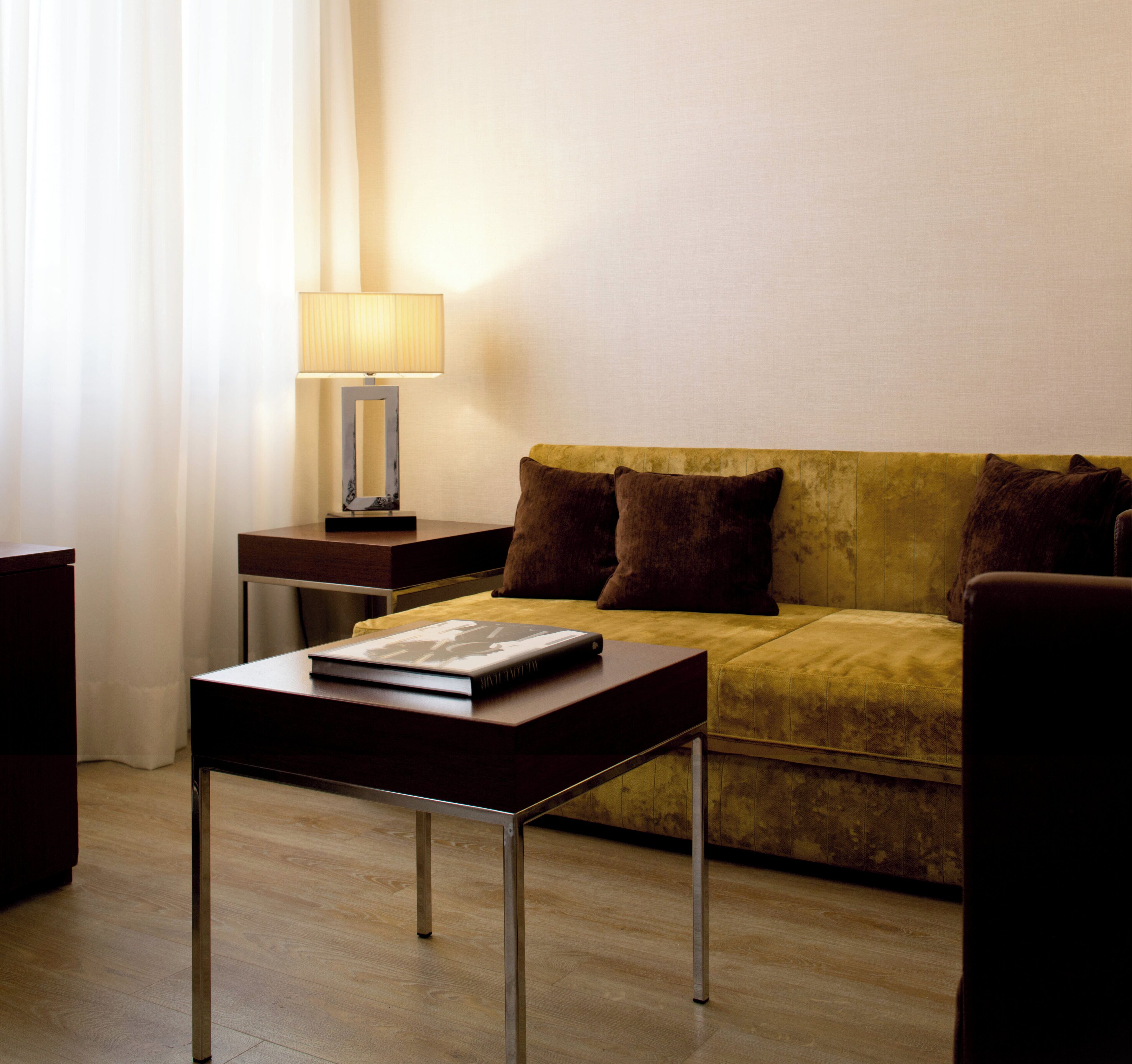 Ein modernes Hotelzimmer mit Holzboden, einem gelben Sofa, einem kleinen Tisch mit einem Buch und einer Lampe auf einem Nachttisch.