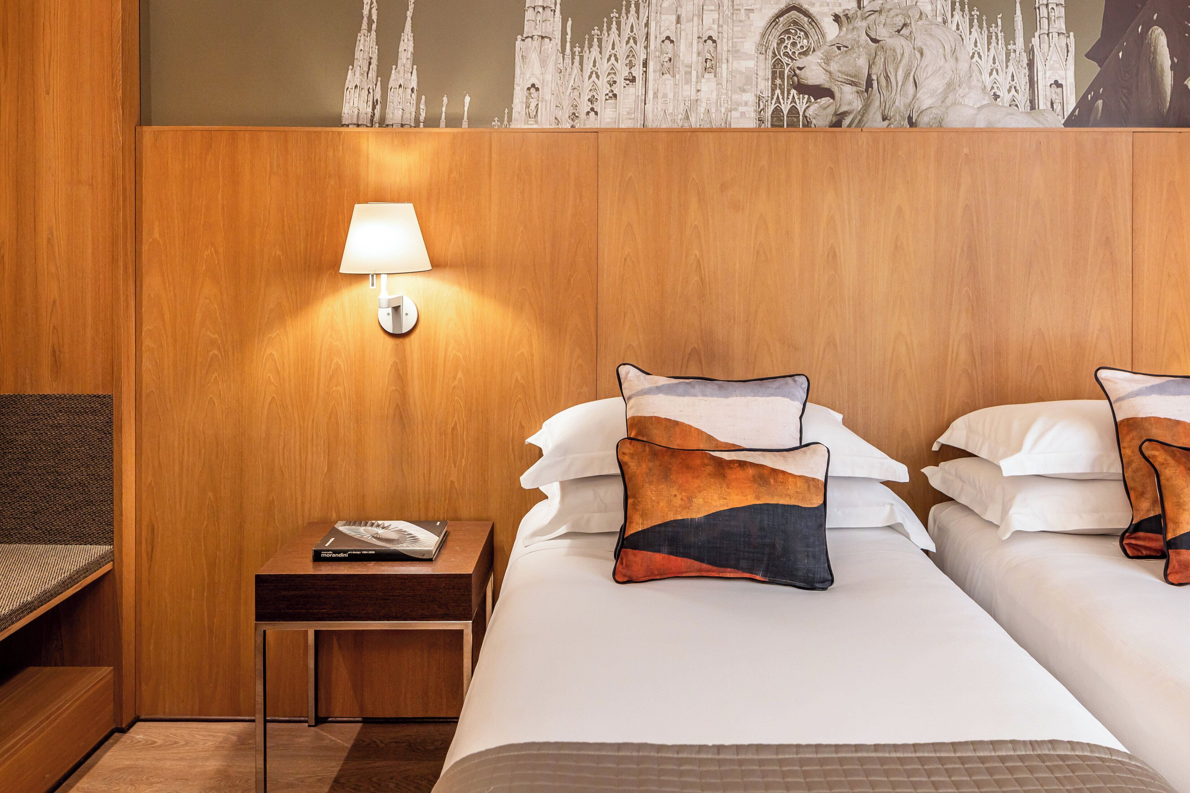 A hotel room with a wooden headboard, a nightstand with a lamp, and two beds with decorative pillows.