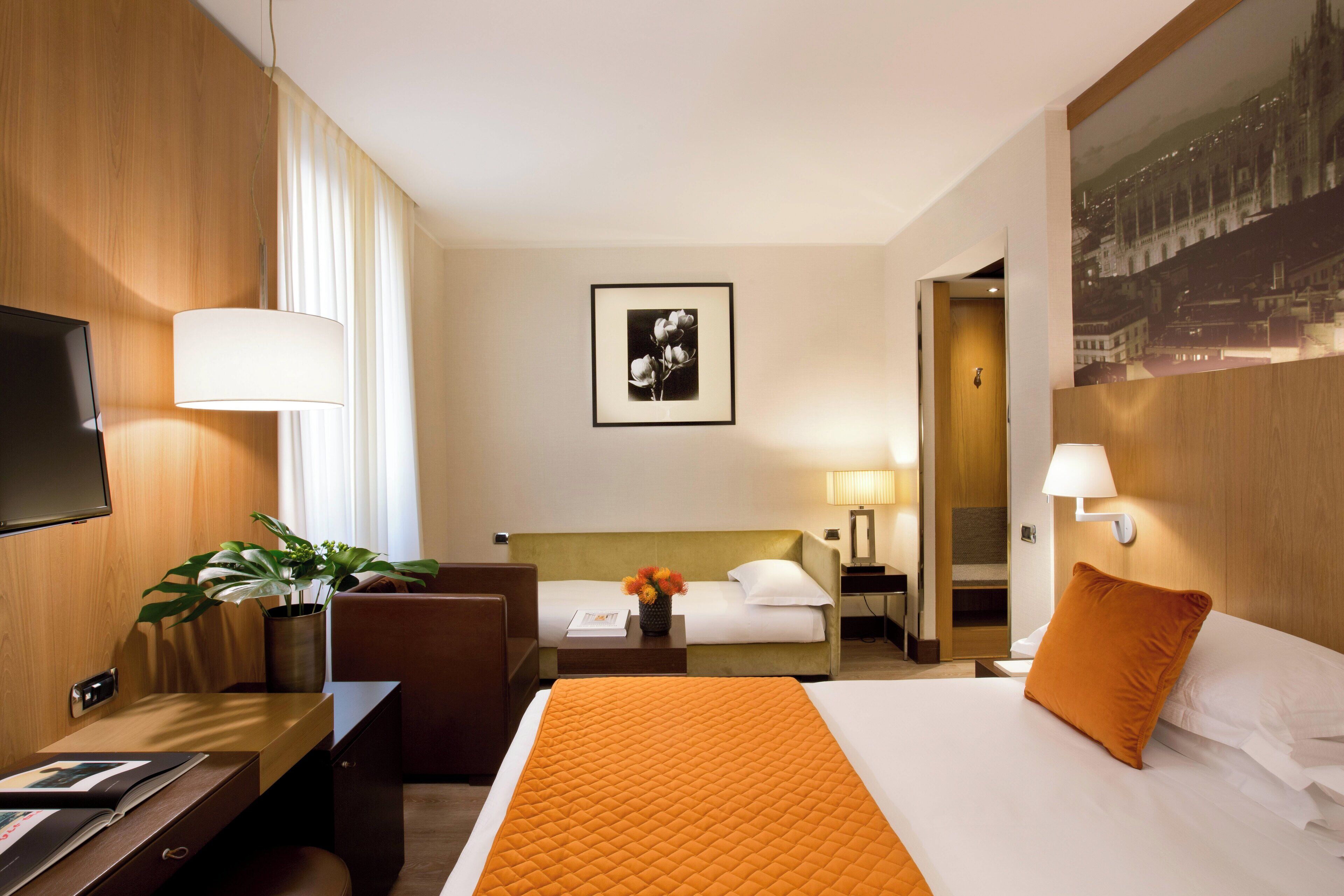 A hotel room with a bed, a desk, a chair, a TV, and a painting on the wall.