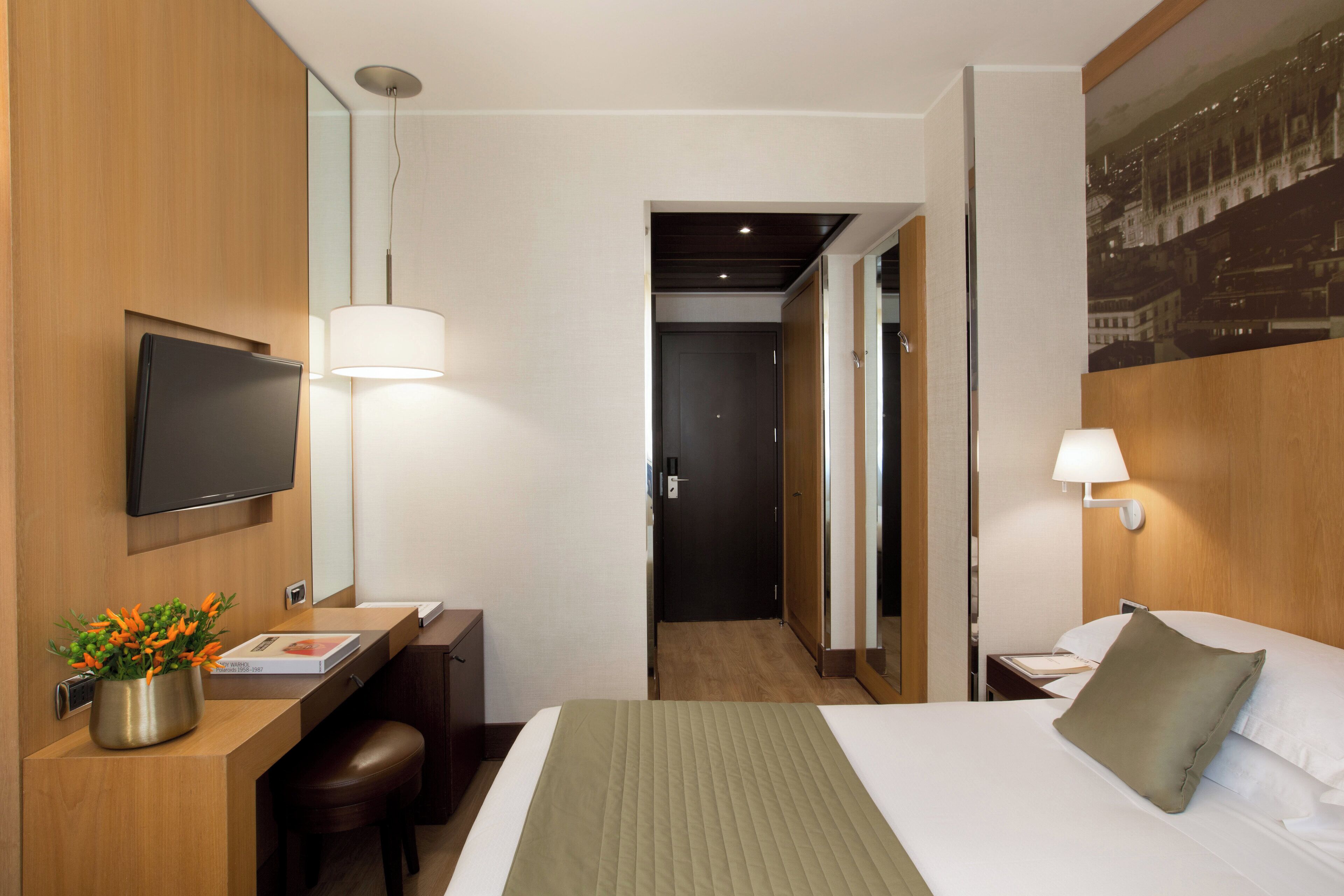 A hotel room with a bed, a desk with a chair, a TV mounted on the wall, and a lamp.