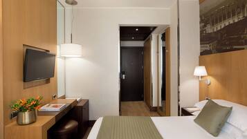 A hotel room with a bed, a desk with a chair, a TV mounted on the wall, and a lamp.