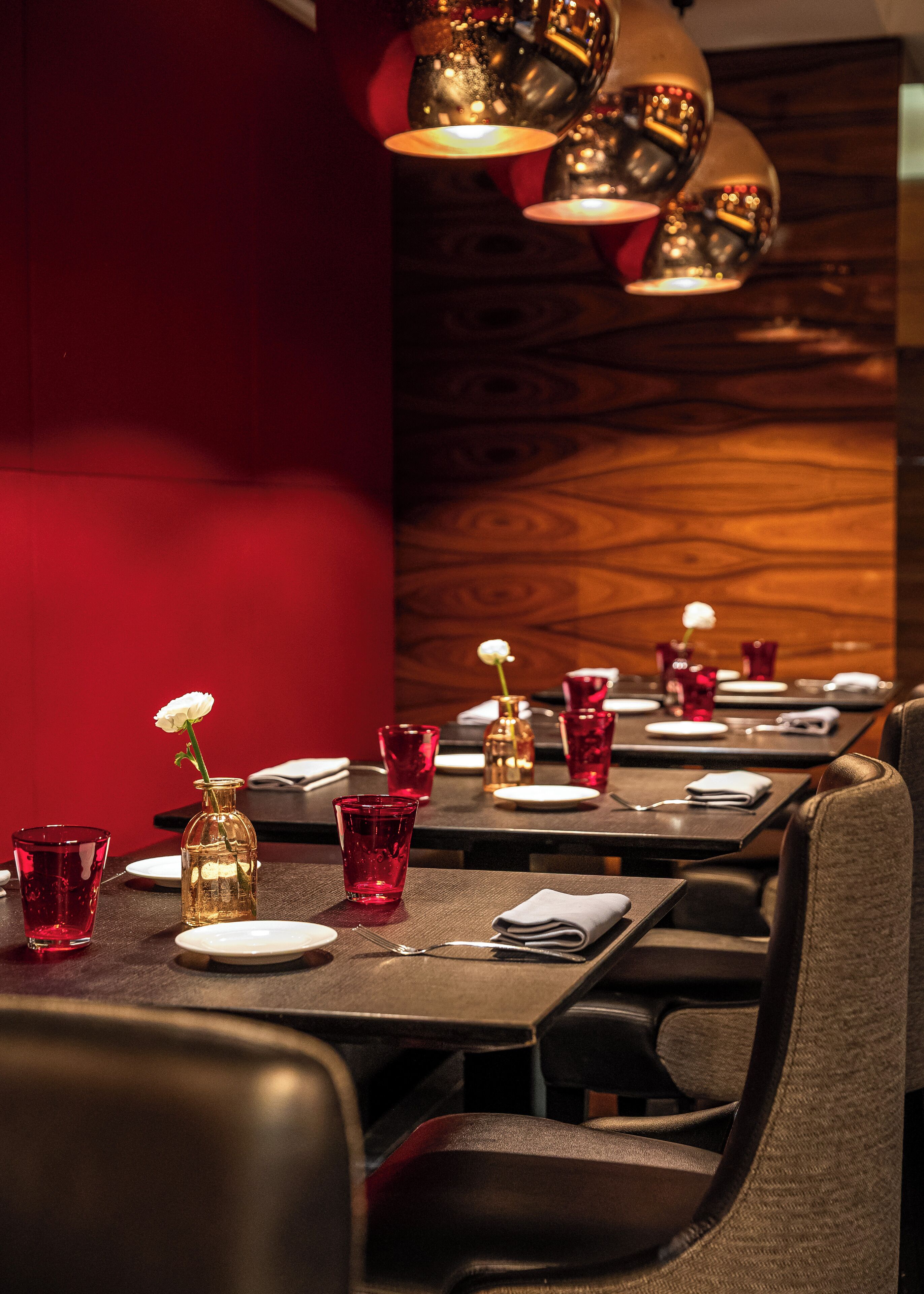 A restaurant setting with tables set for dining, featuring red glassware and a white flower centerpiece.
