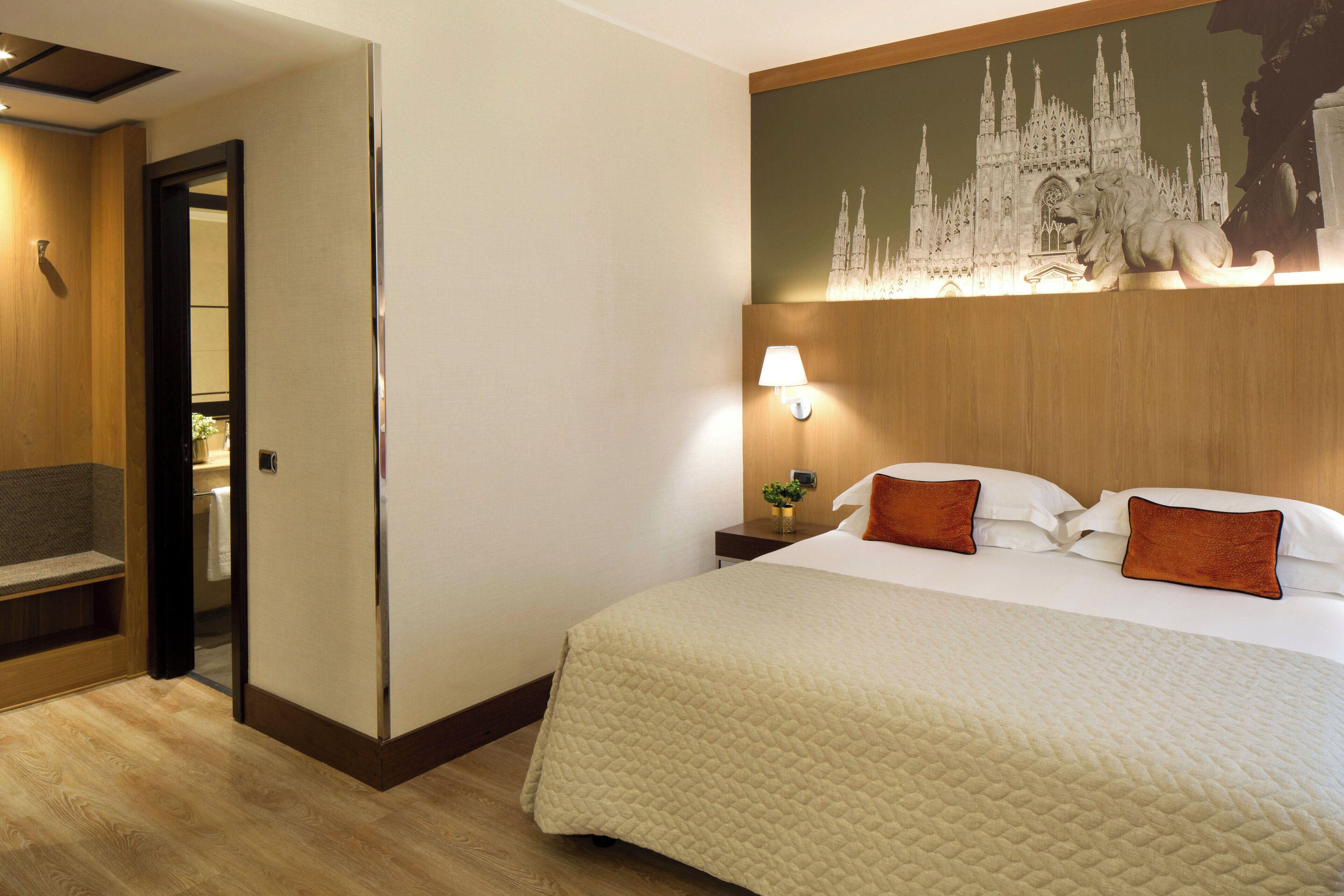 A hotel room with a large bed, a nightstand with a lamp, a wall-mounted artwork of a cathedral, and a wooden bench by the door.