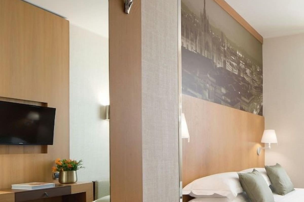 Ein Hotelzimmer mit Bett, Nachttisch, Lampe, Schreibtisch mit Stuhl und Fernseher.