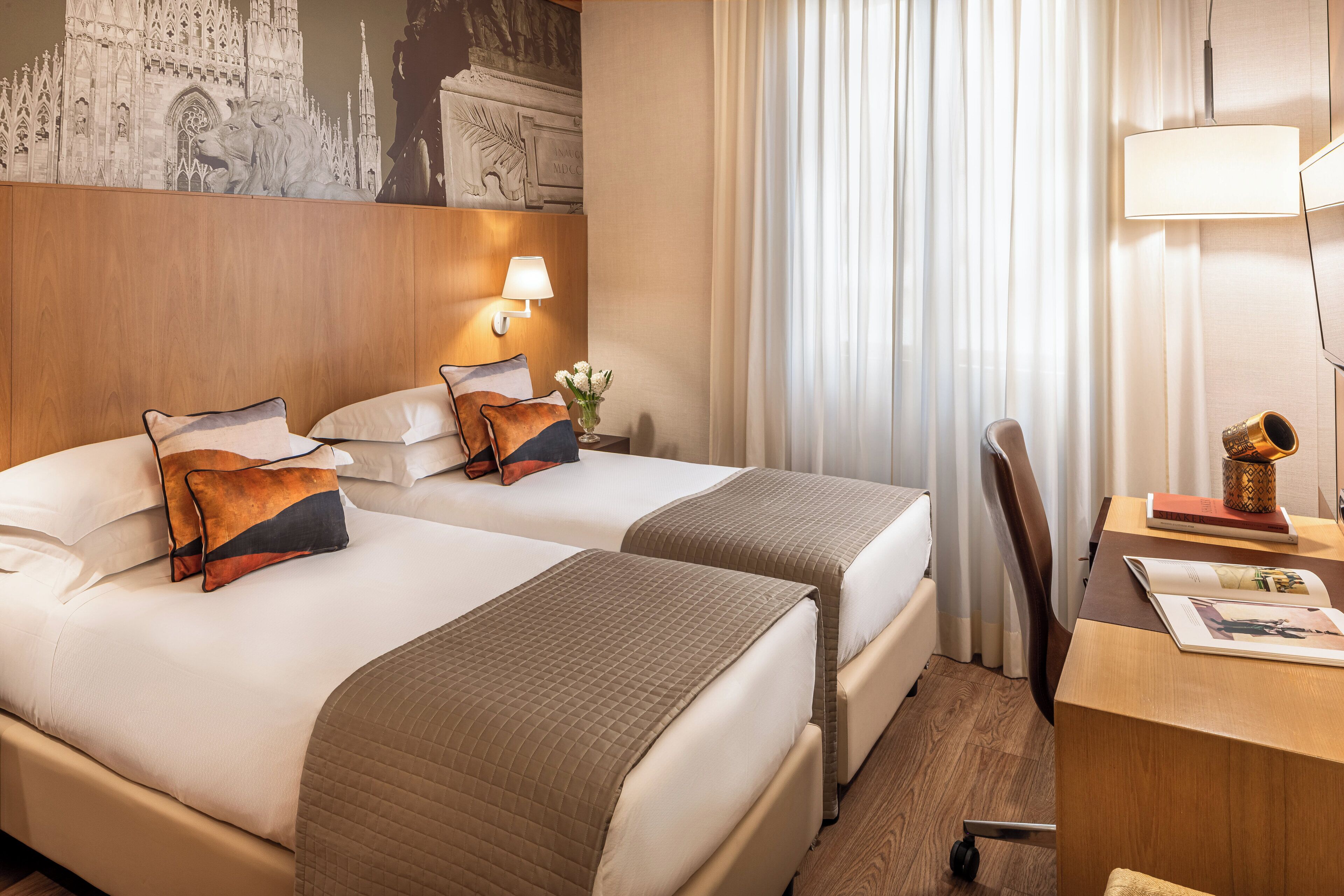 A hotel room with two beds, a desk, a chair, a lamp, and a decorative wall piece.
